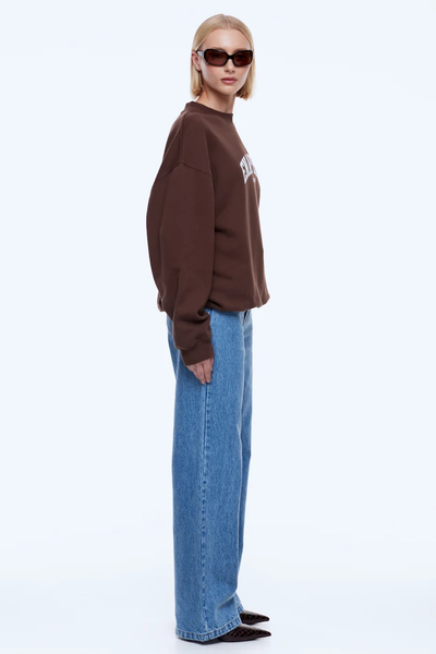 Side profile studio portrait of a model wearing the dark chocolate ENA PELLY Mila Oversized Sweater featuring dropped shoulders, paired with straight-leg denim jeans and dark pointed boots.