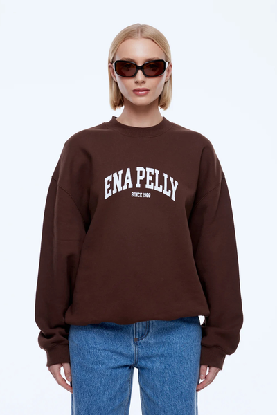 Front half-body view of a model in black sunglasses wearing the chocolate brown ENA PELLY Mila Oversized Sweater showcasing the white arch text chest graphic and rib-trimmed cuffs.
