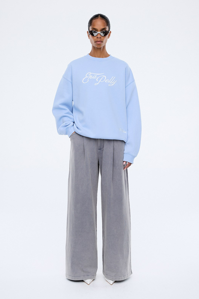 A full-length studio lookbook photograph from the rear, showing the back of the model as she faces away from the camera. The shot highlights the clean, unadorned back panel of the soft blue sweater, its generous, roomy cut, and how the banded hem rests effortlessly over the relaxed grey trousers.