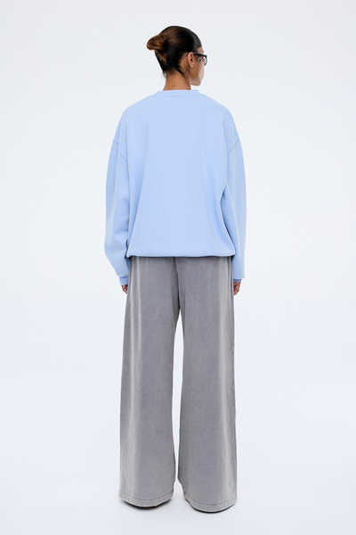 A full-length studio profile photograph capturing the model standing sideways against a plain white backdrop. This angle emphasizes the dramatic slouchy silhouette of the drop sleeves and the extended volume of the premium brushed fleece fabrication through the body.