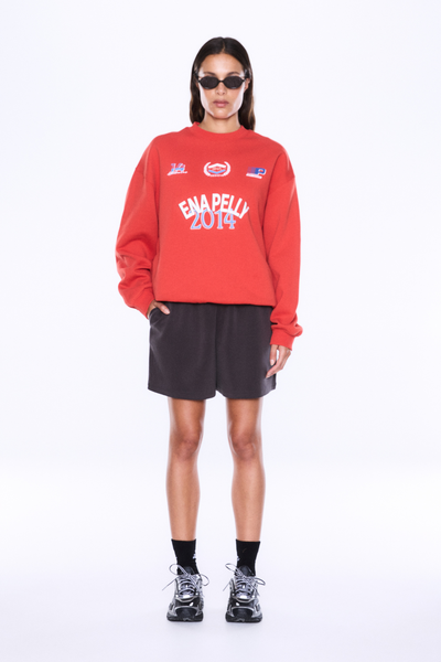 Full-length front studio shot of a model wearing the ENA PELLY Mila Oversized Sweater in Vintage Red with one hand in her pocket, paired with dark casual shorts and sneakers.