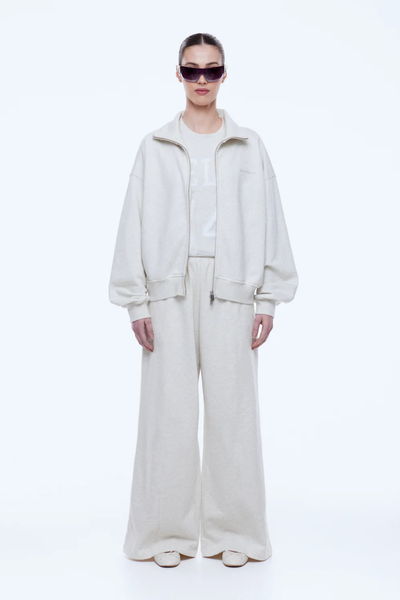 Full-length front view of the ENA PELLY Paris Terry Zip Thru in Oat Marle unzipped over a tonal graphic tee, paired with wide-leg track pants.