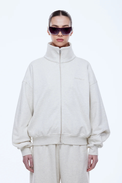 Medium shot of the ENA PELLY Paris Terry Zip Thru in Oat Marle, showing the fully zipped high neck, ribbed cuffs, and subtle tonal branding on the left chest.