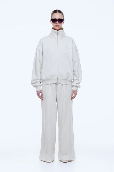 Full-length front shot of a model standing straight in the fully zipped ENA PELLY Paris Terry Zip Thru in Oat Marle and matching wide-leg lounge pants.