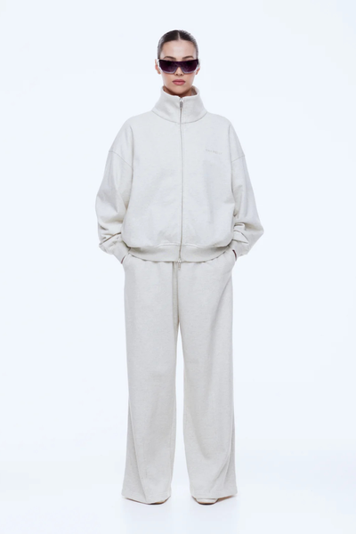 Full-length front studio shot of a model wearing the ENA PELLY Paris Terry Zip Thru in Oat Marle, fully zipped with a high stand collar, hands in pockets, paired with matching wide-leg pants.