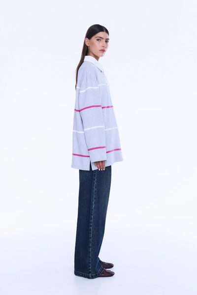 A full-length rear view of the model from behind, showcasing the continuous pattern of the horizontal white and pink stripes across the back of the grey marle polo top, as well as the straight, relaxed hemline over wide-leg dark denim jeans.