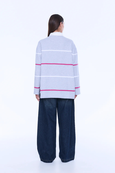 A full-length rear view of a model from behind, showcasing the continuous horizontal pattern of the white and pink sporty stripes across the back of the grey marle polo top. The image highlights the relaxed, long-sleeve silhouette, the solid white contrast collar, and the straight hemline with subtle side splits, paired with dark-wash wide-leg denim jeans.