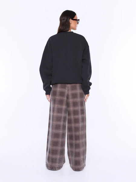 A full-length profile studio shot of a model showcasing the oversized silhouette and effortless drape of the Mila Sweater in Phantom black. The image captures the relaxed drop shoulder cut and the cozy fit of the ribbed cuffs, paired with fluid plaid wide-leg track pants.