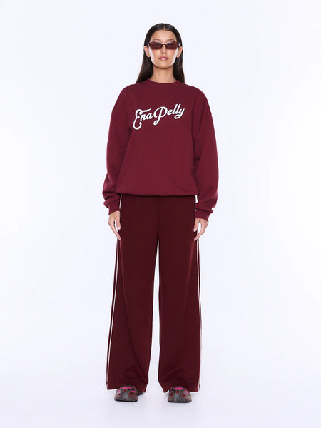 A full-length front studio shot of a model styling the ENA PELLY Mila Oversized Sweater in burgundy. The crewneck sweatshirt showcases a script "Ena Pelly" logo graphic on the chest in vintage white boucle texture with fine black contrast embroidery, paired with matching burgundy wide-leg pants featuring a white side stripe.