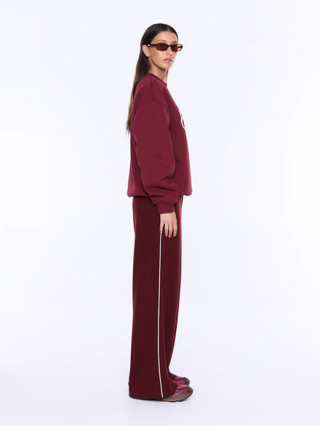 A full-length side profile studio shot of a model wearing the burgundy Mila Oversized Sweater, illustrating the relaxed silhouette and comfortable drape of the brushed fleece fabric. The monochromatic lounge look is completed with fluid wide-leg track pants and tinted sunglasses.
