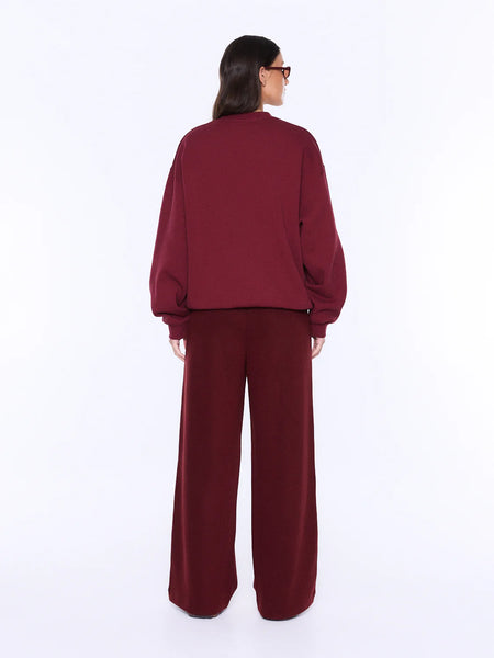 A full-length rear view studio shot of a model wearing the ENA PELLY Mila Oversized Sweater in burgundy. The image emphasizes the minimalist, clean back construction of the cotton-blend brushed fleece sweatshirt and its loose, oversized fit over the shoulders and waist.