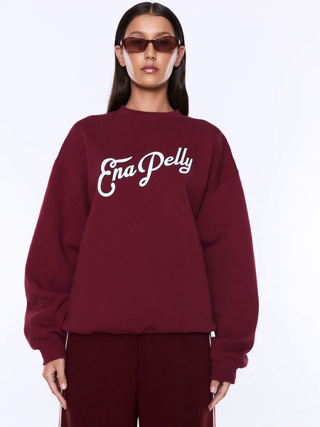 A close-up studio portrait capturing the front details of the ENA PELLY Mila Oversized Sweater in burgundy. The shot highlights the clean crew neckline, relaxed drop shoulders, and the textured vintage white boucle script brand name with fine black embroidered outlines on the chest.
