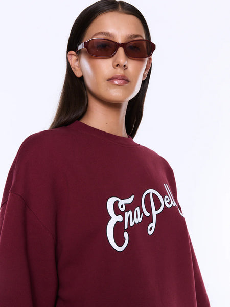 A macro detail shot focusing on the chest of the burgundy Mila Sweater, highlighting the unique tactile quality of the exclusive Ena Pelly script graphic. The close-up showcases the plush texture of the vintage white boucle fabric against the smooth burgundy brushed fleece.