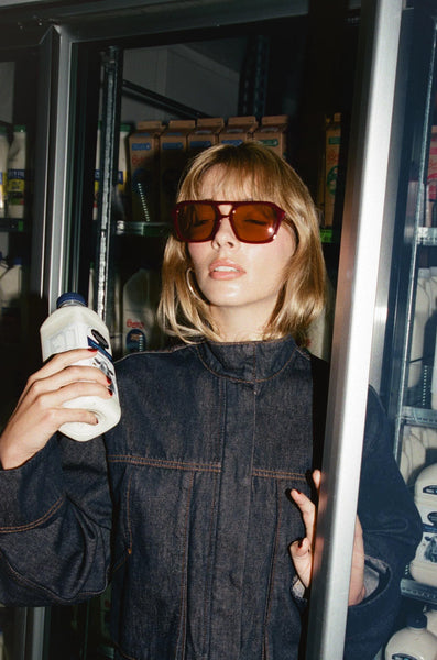A retro-themed lifestyle campaign photograph of a blonde model with a fringe inside a grocery store setting, holding a bottle of milk near an open glass refrigerator door. She is wearing the oversized milky berry aviator sunglasses paired with a dark denim utility jacket, evoking an authentic 1970s vintage aesthetic.