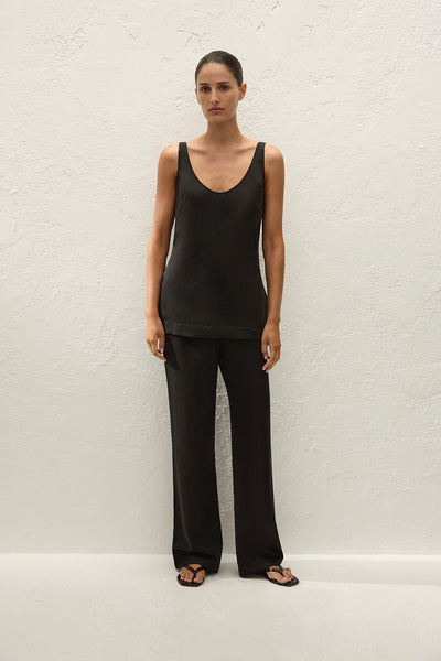 Full-length studio front view of a model wearing the black sleeveless Varina scoop-neck top styled with matching black wide-leg tailored trousers and black thong sandals against a textured beige wall.