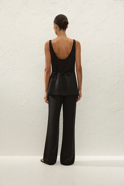 Full-length studio rear view of a model wearing a black scoop-back sleeveless top featuring a delicate back waist tie cinch paired with black wide-leg pants.