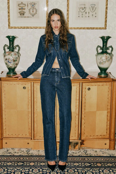 Editorial-style portrait of a model with long wavy brown hair posing in front of an ornate wooden cabinet flanked by two large green ceramic vases. She is wearing the Neuw Freja Straight jeans paired with a matching dark indigo denim shirt, showcasing a classic double-denim look.
