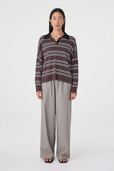 A full-length front studio shot of a model wearing the Arcaa Movement Rayne Knit Jersey in Espresso. The image displays the oversized polo-style silhouette, custom striped print, contrast black collar, and casual styling with gray wide-leg tailoring against a plain white background.
