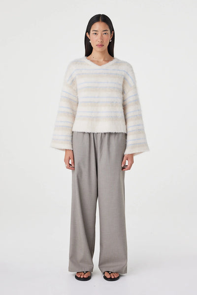 A full-length front studio shot of a model presenting the Arcaa Movement Lucca Sweater in Taupe and Blue Stripe. The image clearly showcases the soft, fluffy knit texture, the relaxed V-neckline silhouette, dropped shoulders, and wide sleeves, styled with loose gray wide-leg trousers.