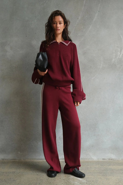 A full-length front studio shot of a model wearing the high-rise Arcaa Movement Emery Pant in Merlot and Cream. The image highlights the straight leg silhouette, paired with a matching merlot knit pullover and black sandals against a plain white background.