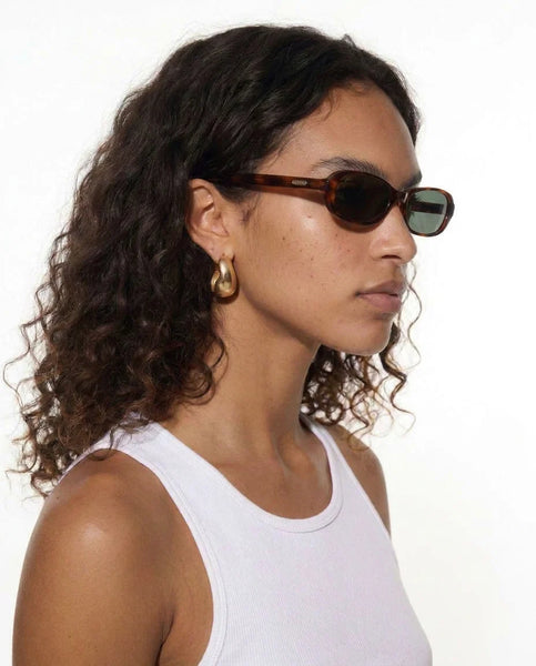 Close-up profile portrait of a dark-haired female model with curly hair wearing the Sito Shades Roux sunglasses in Classic Tort acetate paired with chunky gold hoop earrings.