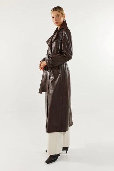 A full-length front studio image of a model wearing the chocolate brown faux leather trench coat styled open and unbuttoned over a black scoop-neck top and cream wide-leg trousers, completed with black pointed-toe boots and rectangular sunglasses.