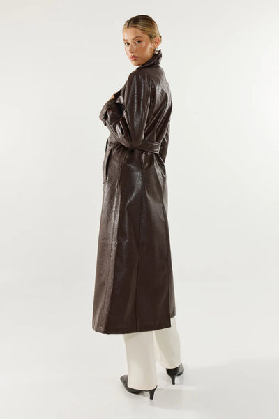 A full-length rear profile studio shot of the model in the rich chocolate long-line trench coat, demonstrating the relaxed fit silhouette and supple drape of the textured faux leather fabrication from behind.