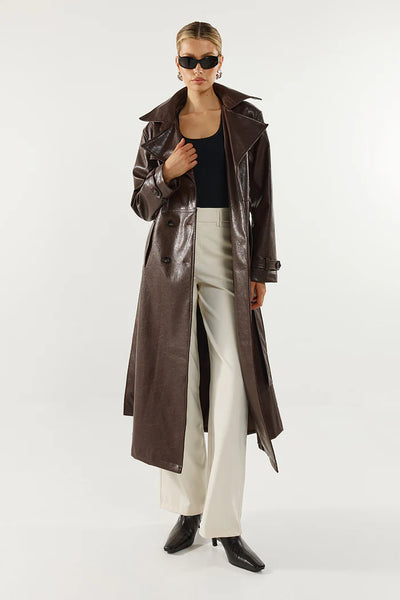 A side-angle studio product shot of the model showcasing the side-profile details of the chocolate brown double-breasted coat, emphasizing the extended collar, cuff straps, and wrapped waist tie.