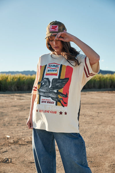 A candid close-up lifestyle photograph focusing on the front of the beige cotton tee worn by a model out in nature, showcasing the bright green, red, and cream tones of the detailed vintage-style rodeo print across the chest.