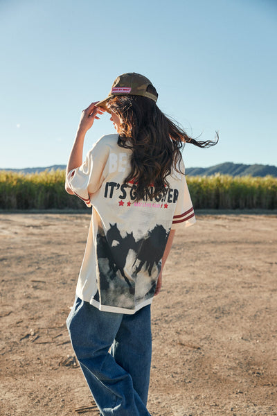 A full-length studio product shot from the back on a model wearing the beige oversized graphic tee as a dress, highlighting the large, striking back graphic featuring a dark silhouette illustration of running wild horses topped with bold retro typography.