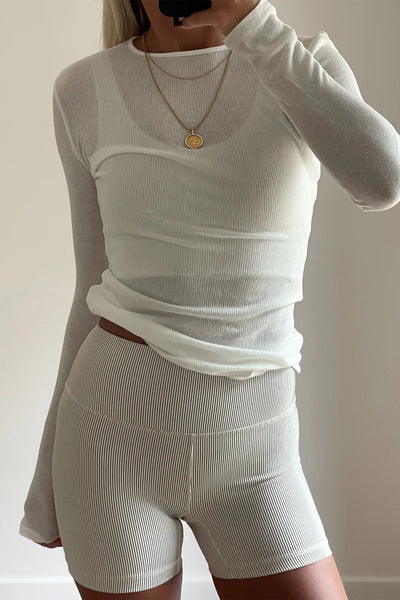 Close-up selfie shot of a model wearing the semi-sheer white Zulu & Zephyr Rib Hemp Jersey Long Sleeve Top styled with a gold pendant necklace and ribbed shorts.