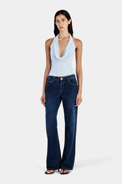 Studio front view of a model wearing a sky blue jersey halter cowl neck top paired with dark blue denim jeans.