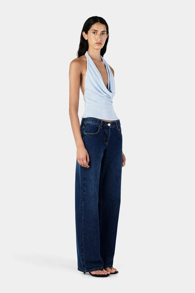 Studio three-quarter front view of a model wearing a sky blue jersey cowl neck halter top paired with dark wash jeans.