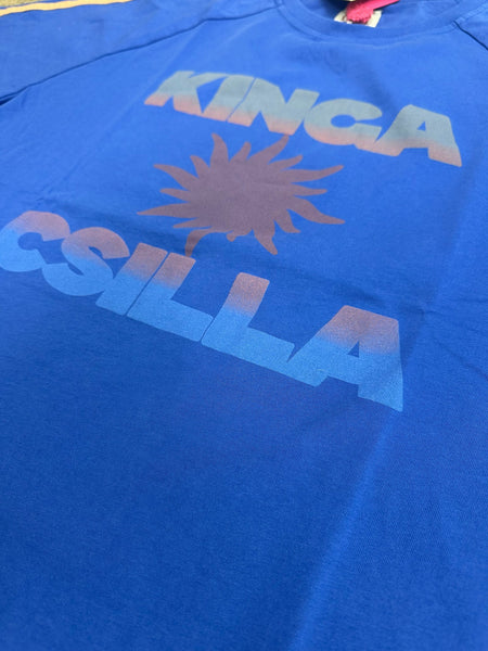 A flat-lay, close-up shot focusing on the center graphic of the bright Nuwa Blue cotton jersey Sunset Tee. The image showcases the screen-printed "KINGA CSILLA" branding surrounding a sunburst motif, rendered in a faded gradient of coral and sky blue hues.