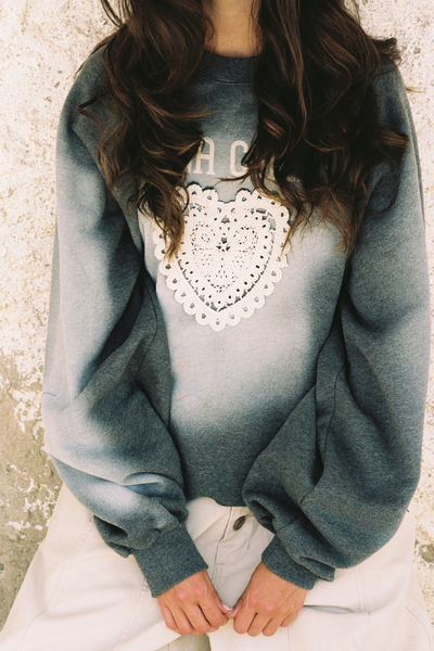 A medium shot of the model walking outdoors past a stone archway, wearing the slouchy, relaxed-fit jumper paired with off-white, wide-leg trousers. The image highlights the loose, softly structured drape of the 100% cotton terry cloth and the screen-printed "Kinga Csilla" branding curved above the lace heart.