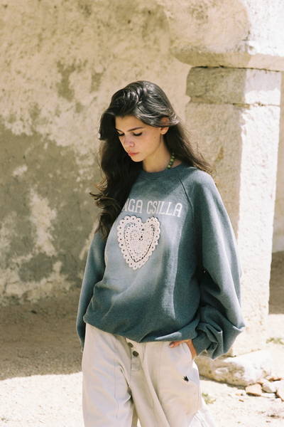 A profile shot of the model looking off-camera against a rustic wall, holding her hands together. Her arm is lifted to display the rectangular tan brand patch stitched onto the right sleeve, which features embroidered floral motifs and text.