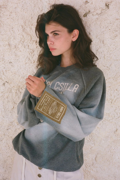 A studio lookbook photograph of a model with her hair in a bun, standing against a solid blue backdrop. She has her hands on her head, showcasing the oversized, slouchy fit of the raglan-sleeve pullover and its vintage-wash look, styled over a sheer, dark lace asymmetrical skirt.