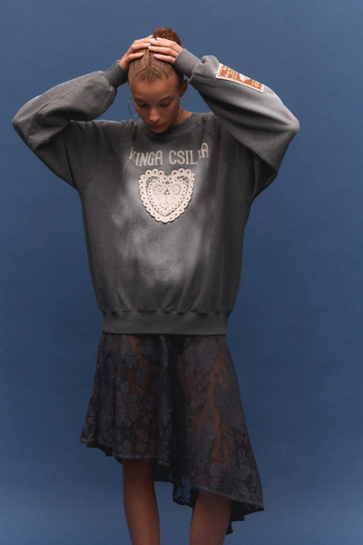 A full-length studio portrait of the model against a blue background, standing with her hands loosely clasped in front. This shot highlights the complete outfit silhouette, showing the jumper's ribbed hemline resting easily over the sheer lace skirt, finished with studded brown mule slides.