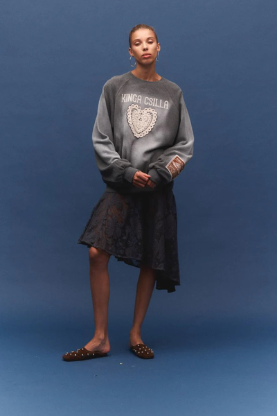 A front-on, medium portrait of the brunette model leaning directly against a neutral-toned plaster wall. Her relaxed posture emphasizes the cozy, oversized proportions and dropped shape of the natural-fiber knit pullover.