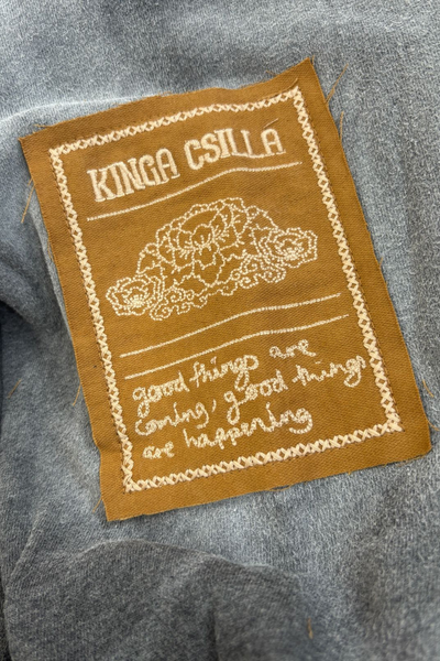 A macro detail shot focusing on the rectangular, golden-brown woven canvas patch on the right sleeve. The patch features raw edges and white embroidered detail displaying the "Kinga Csilla" logo, a central peony floral design, and the cursive script: "good things are coming, good things are happening.