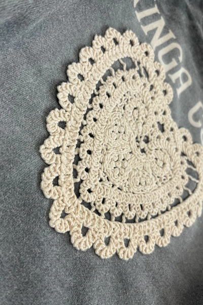 A texture-focused macro shot showing the intricate craftsmanship of the cream crochet 'Day Hearts' medallion stitched onto the charcoal marl fabric. The image clearly displays the looped knit patterns, scalloped lace trim, and the soft, heathered loops of the premium cotton base.