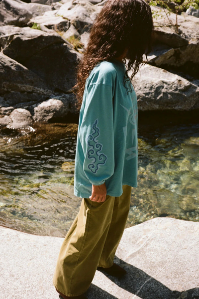 Outdoor lifestyle side profile shot of a model wearing the long-sleeve Kinga Csilla Paper Sun Roses Tee in Blue, highlighting the cloud motif screen-printed along the sleeve.