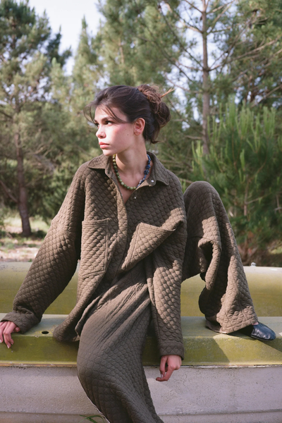 An outdoor over-the-shoulder shot of the model looking away from the camera, highlighting the back of the shirt. This view showcases the curved shirt-tail hem and the bold, black hand-painted graphic art design positioned prominently across the center back of the textured quilted fabrication.