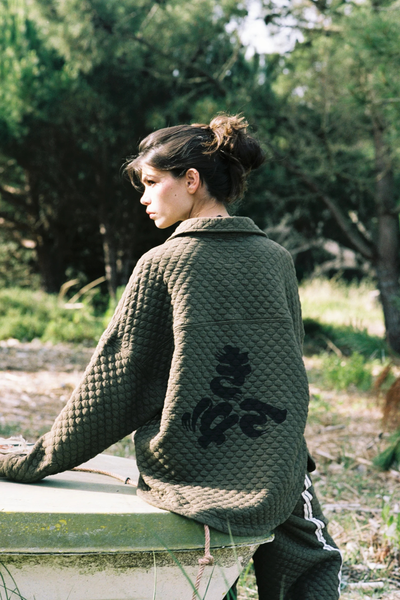 A wide, low-angle outdoor campaign photograph featuring the model standing atop a massive fallen tree trunk in a pine forest. She wears the full quilted khaki set, emphasizing how the slightly boxy shirt and matching wide-leg trousers drape naturally in a outdoor environment.