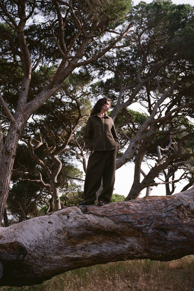 A medium side-profile campaign photograph of the brunette model set against a soft coastal background with water. The shot captures the relaxed, generous fit through the shoulders and arms, as well as the black graphic detailing painted on the textured back panel.
