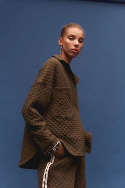 A close-up studio lookbook portrait of a model with short hair looking over her shoulder against a solid blue backdrop. She wears the oversized khaki quilted shirt, showing off the detailed bubble stitching on the dropped sleeves and the clean lines of the front pocket.