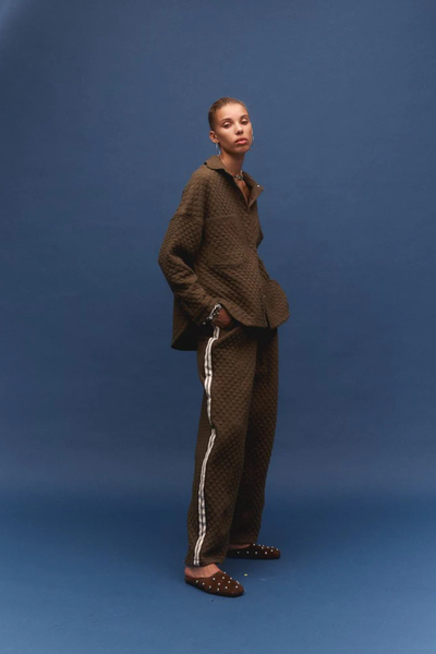 A full-length studio lookbook image of the model against a blue background, standing in a relaxed side profile. She is wearing the Sabbia quilted shirt paired with matching khaki track-style pants featuring a white side stripe, completed with dark brown studded mule slides.