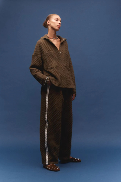 A three-quarter studio portrait of the model standing against a blue background, looking downward. The image displays the slightly unbuttoned neckline of the relaxed utility shirt, showcasing its texture, weight, and casual structure when worn as a standalone piece.