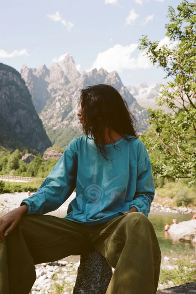 Outdoor lifestyle photo of a model wearing the relaxed-fit long-sleeve Kinga Csilla Paper Sun Roses Tee in Blue with olive wide-leg trousers, seated against a mountain stream landscape background.