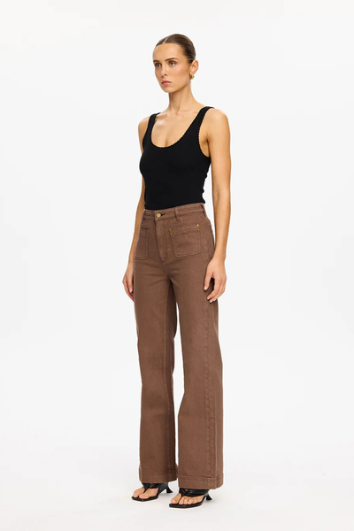 A full-length, three-quarter studio profile shot of a model standing at an angle against a clean white background. She is wearing the espresso brown rigid denim Dominique Jean, paired with a tucked-in scoop-neck black tank top and black leather thong-style heeled sandals, highlighting the fitted silhouette through the waist and hip.